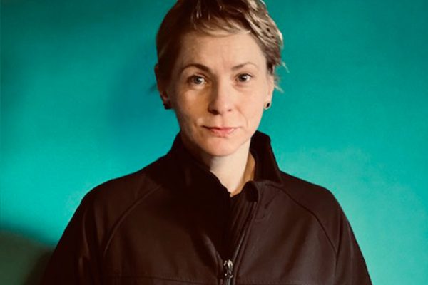 A person with short hair stands against a teal background wearing a black jacket with "Wellways" and "StandBy Support" logos. They have a calm expression.