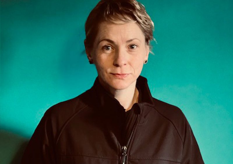A person with short hair stands against a teal background wearing a black jacket with "Wellways" and "StandBy Support" logos. They have a calm expression.