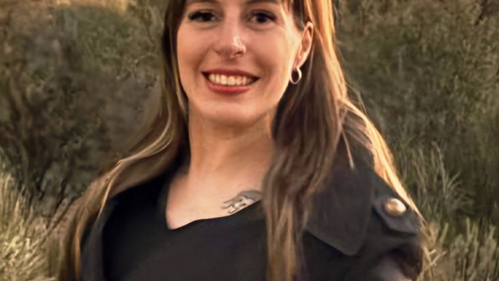 A smiling woman in a black coat stands in front of desert cliffs at sunset. Warm tones create a serene, joyful atmosphere with natural beauty.