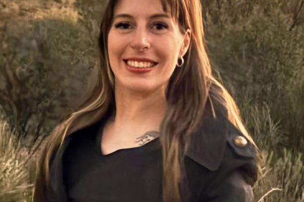 A smiling woman in a black coat stands in front of desert cliffs at sunset. Warm tones create a serene, joyful atmosphere with natural beauty.